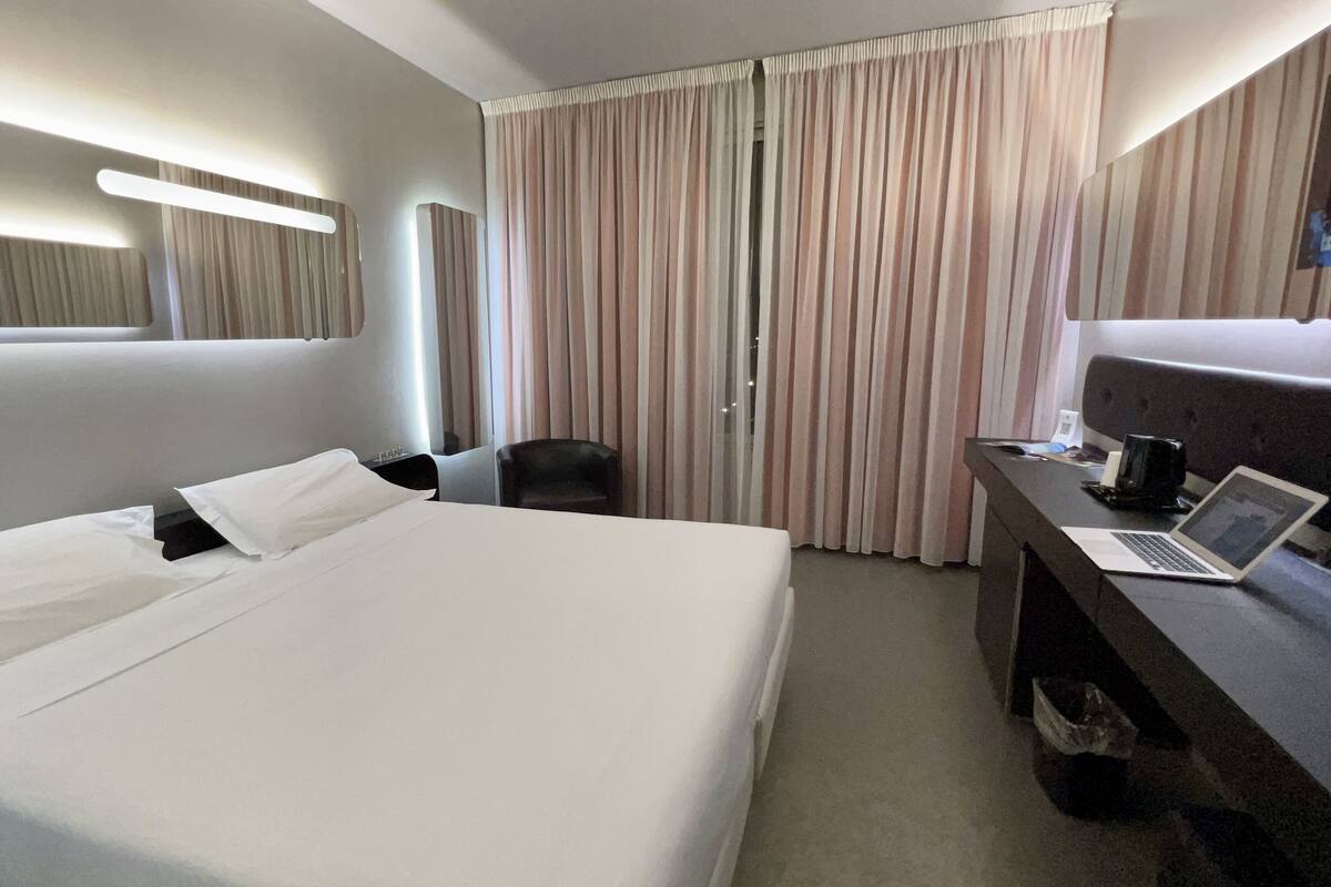 A hotel room with a large bed, a desk with a laptop, a chair, and a window with curtains.