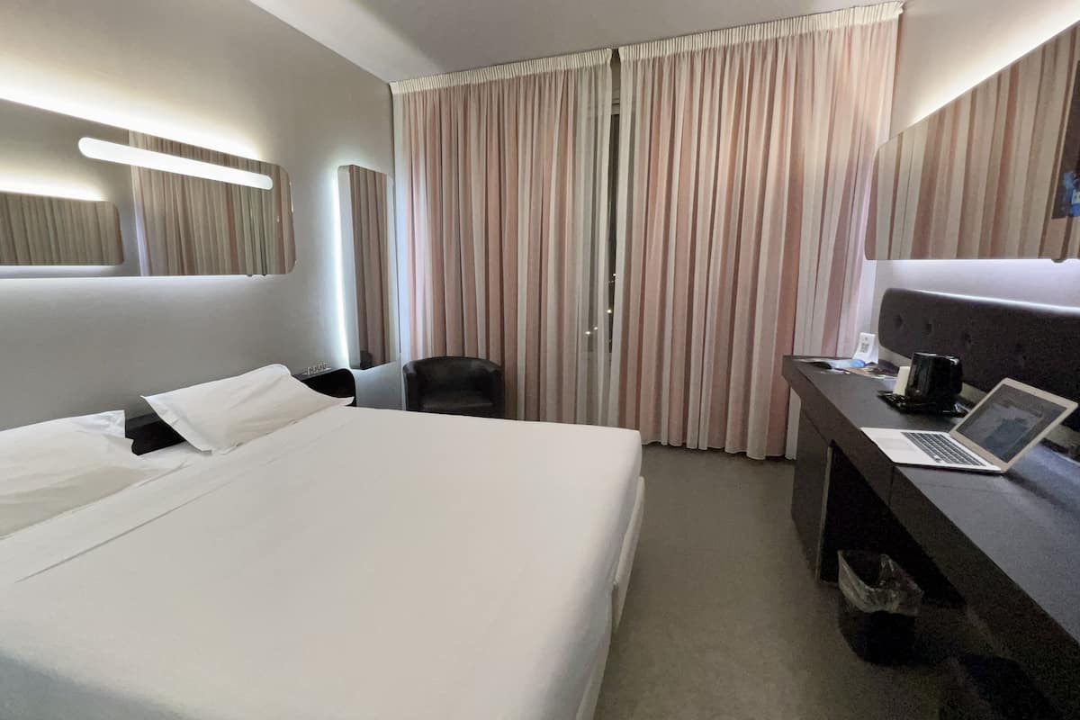 A hotel room with a large bed, a desk with a laptop, a chair, and a window with curtains.