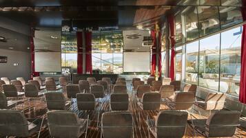A conference room with rows of chairs, large windows with red curtains, and a wooden floor.