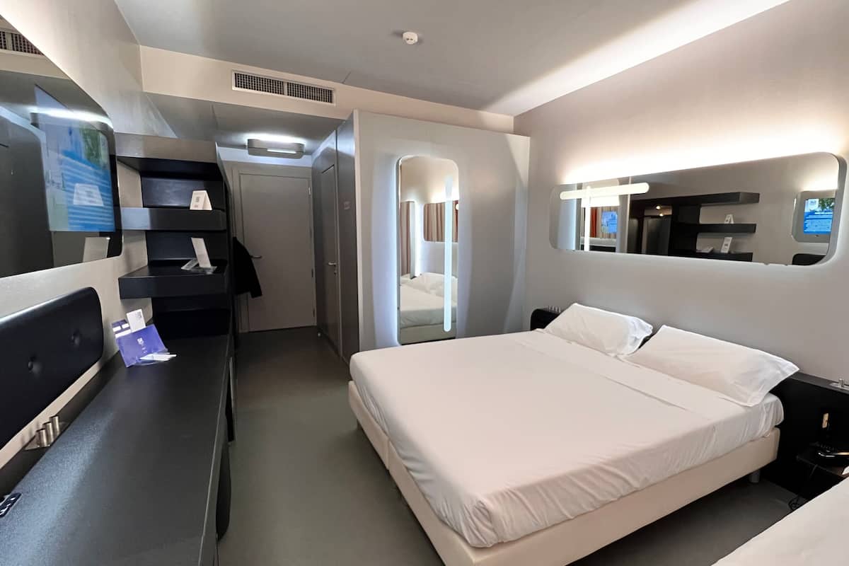 A hotel room with a bed, bedside tables, a desk with a computer, a mirror, and a door leading to a bathroom.
