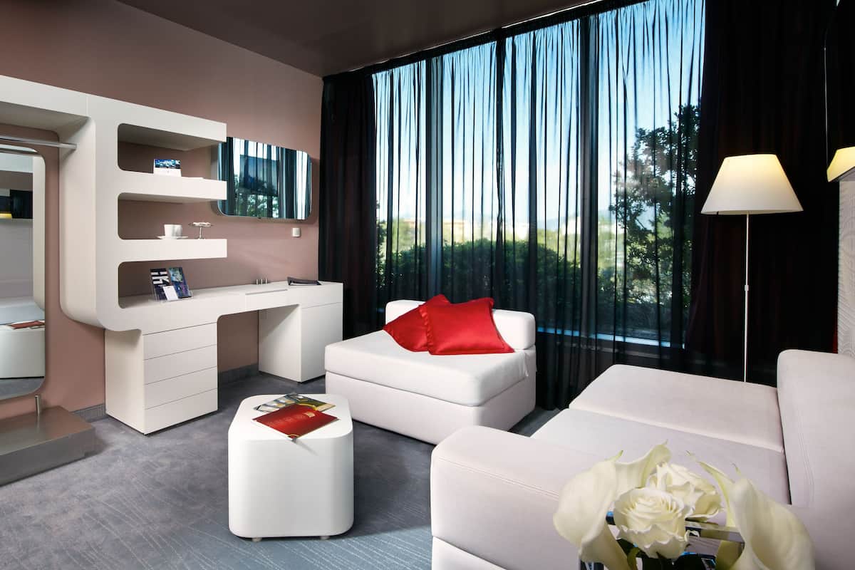 A modern hotel room with a white sofa, a red pillow, a white ottoman, a desk with a mirror, and large windows offering a view of greenery.