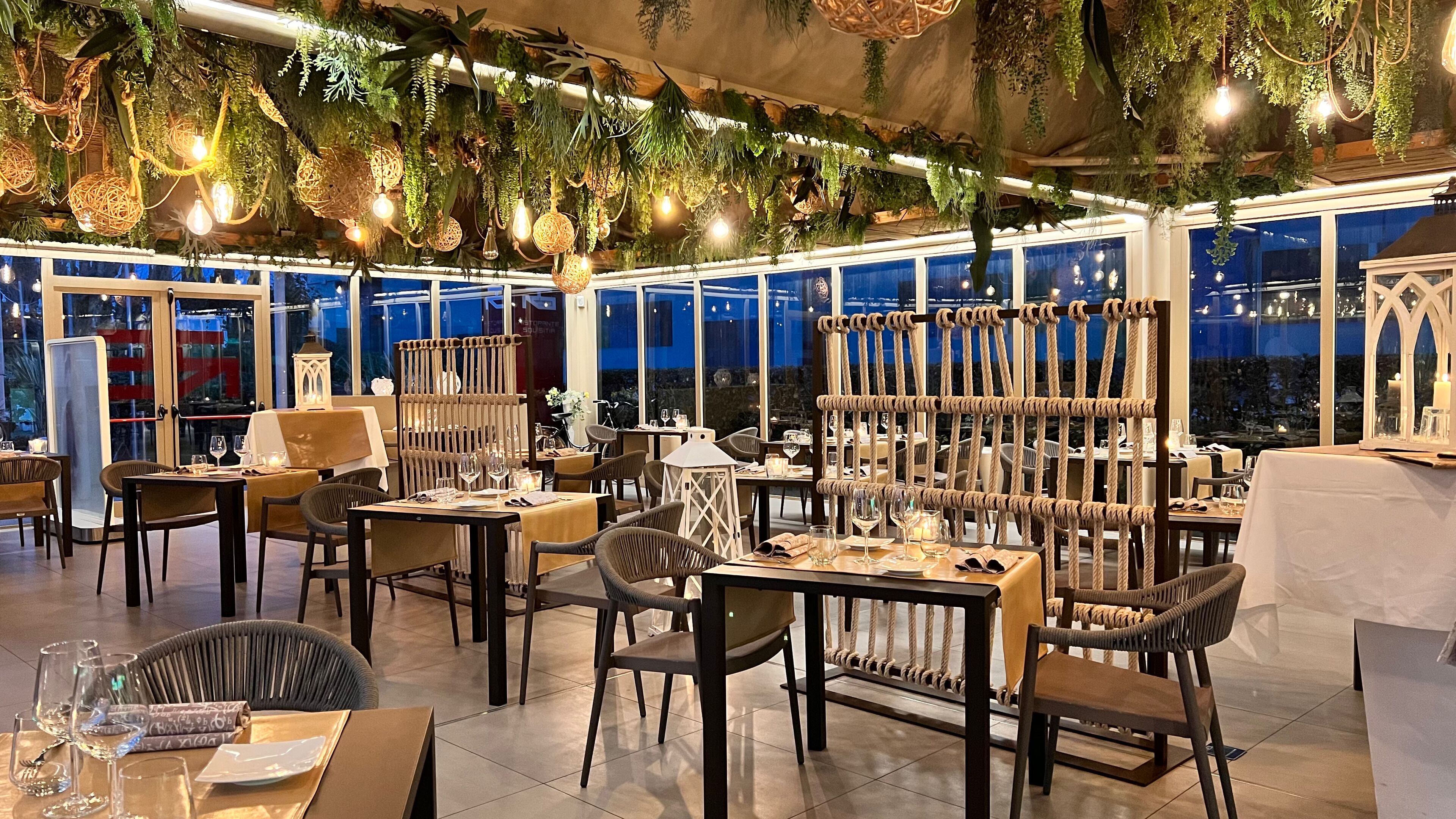 Um restaurante com mesas, cadeiras, plantas penduradas e luzes de Natal.