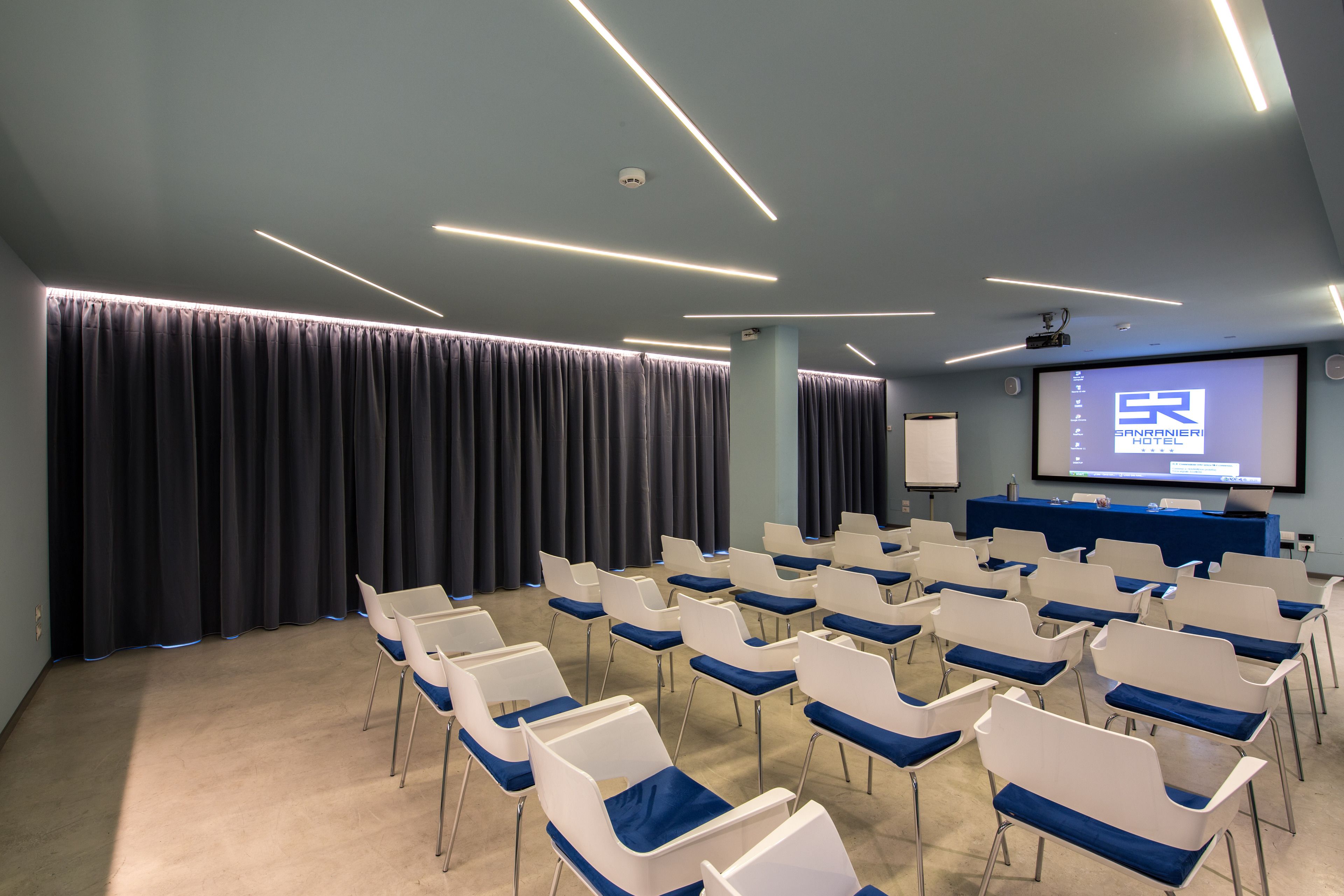 A conference room with rows of white chairs, a projection screen displaying a hotel name, and a blue carpeted floor.