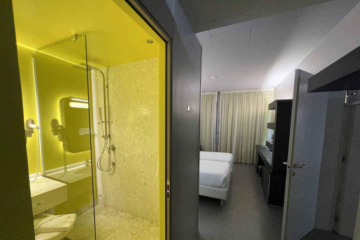 A modern hotel room with a glass-enclosed shower, a separate bathroom, a bed, a desk, and a mirror.