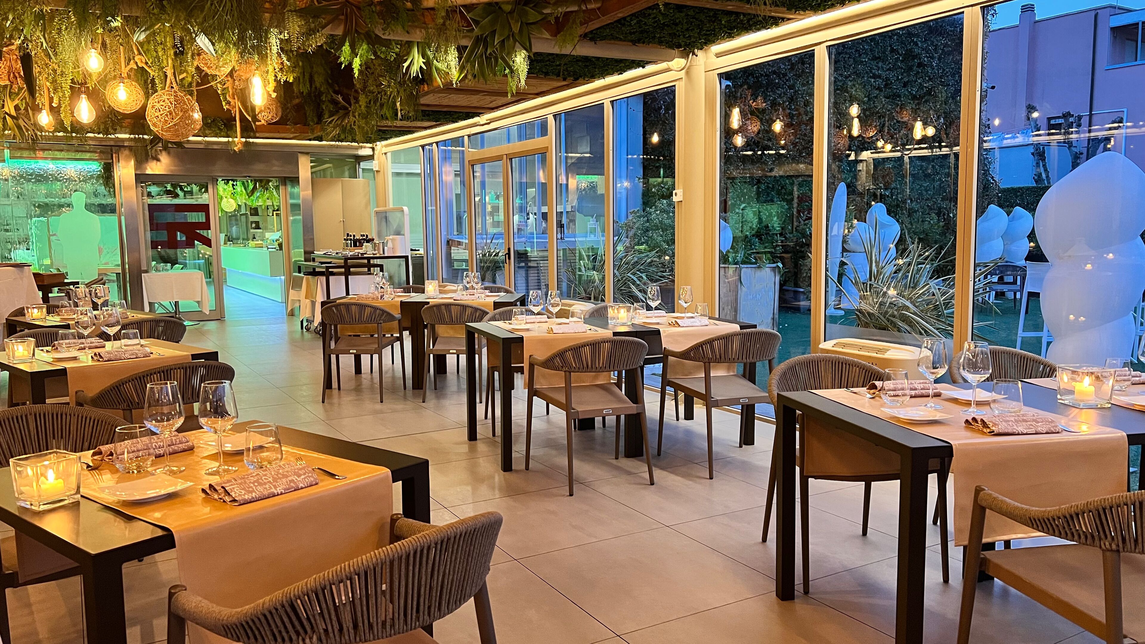 Un restaurante con varias mesas dispuestas para comer, ventanas amplias y plantas colgantes.