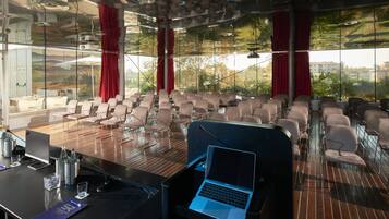 Ein Konferenzraum mit Reihen von Stühlen, einem Laptop auf einem Ständer und einer Glaswand mit Blick auf Gebäude und Bäume.