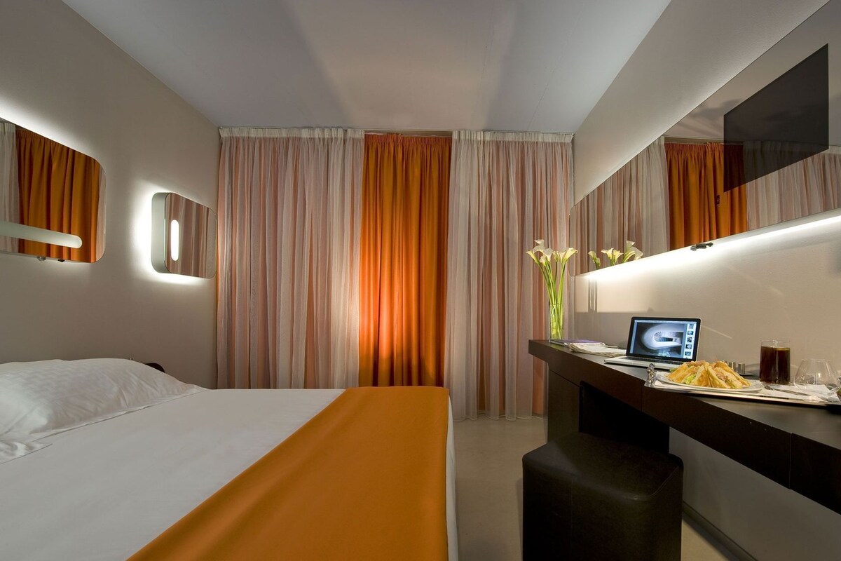 Una habitación de hotel con una cama, un escritorio con una computadora portátil, un espejo y un florero con flores.