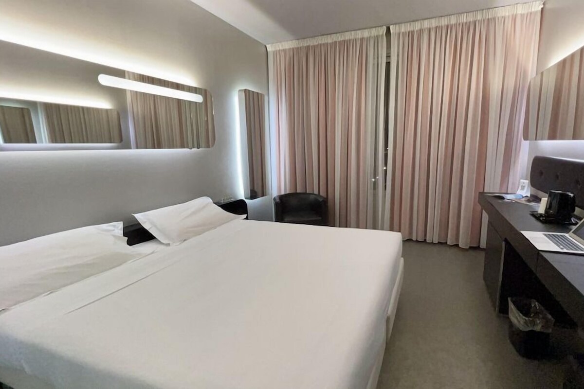 Habitación de hotel con una cama grande, un escritorio con una computadora portátil, una silla y una ventana con cortinas.