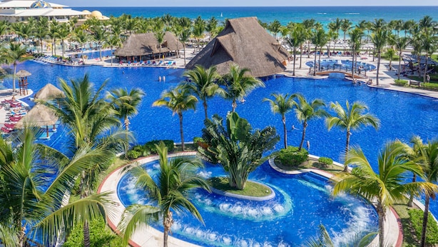 Un resort con una amplia piscina central, palmeras y un edificio con techo de paja.