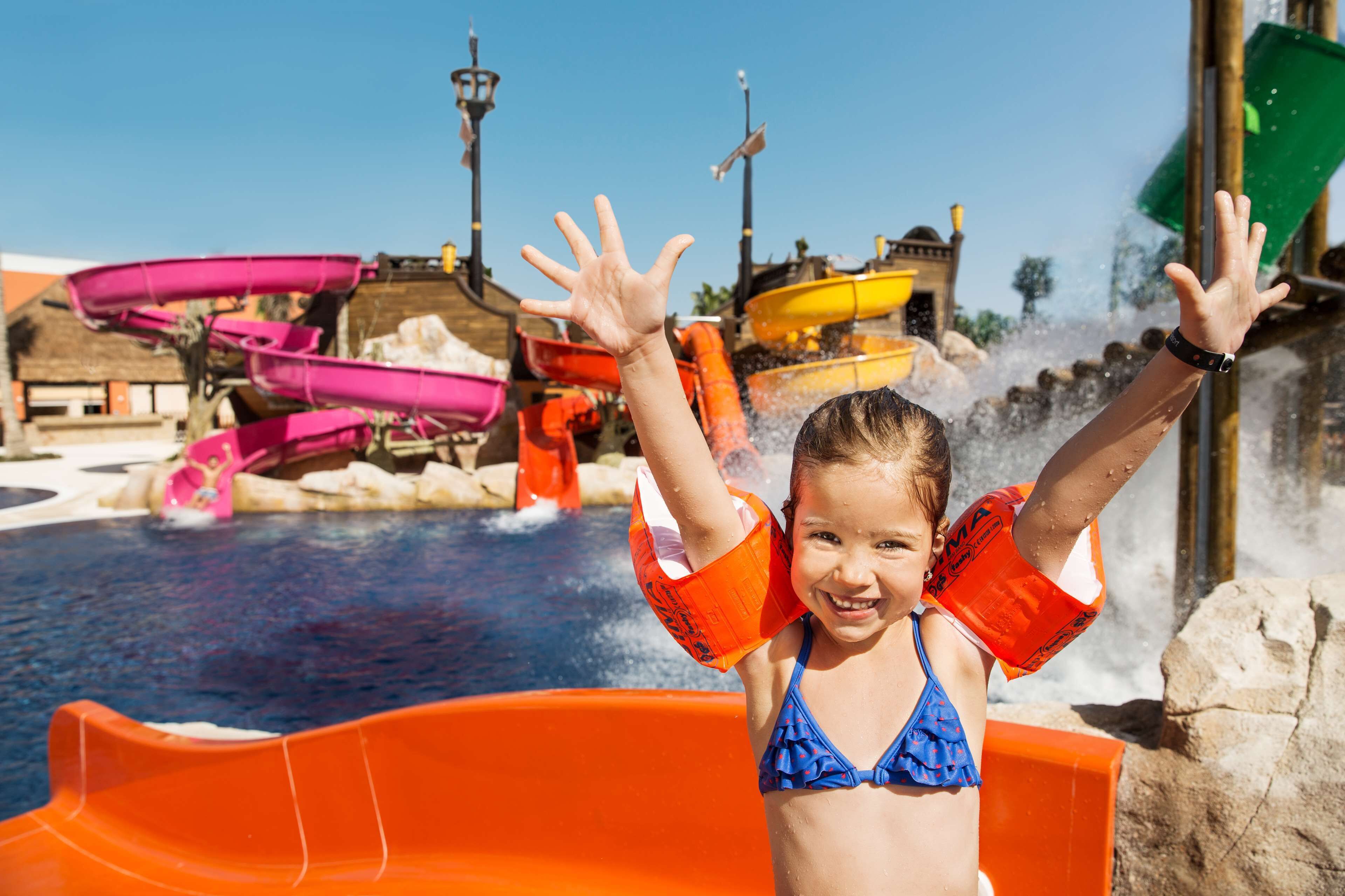 Un enfant, vêtu d’un maillot de bain bleu et portant un gilet de sauvetage orange, se tient dans un toboggan aquatique orange, dans un parc aquatique.