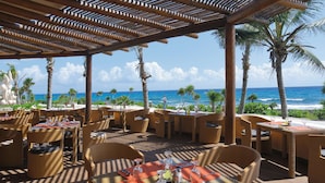 Un espacio de comedor al aire libre con mobiliario de mimbre y mesas dispuestas para comer, con vistas a una playa con palmeras y el océano.