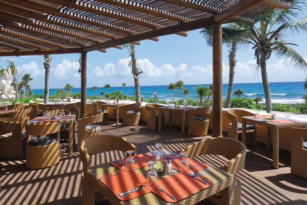 Un espacio de comedor al aire libre con mobiliario de mimbre y mesas dispuestas para comer, con vistas a una playa con palmeras y el océano.