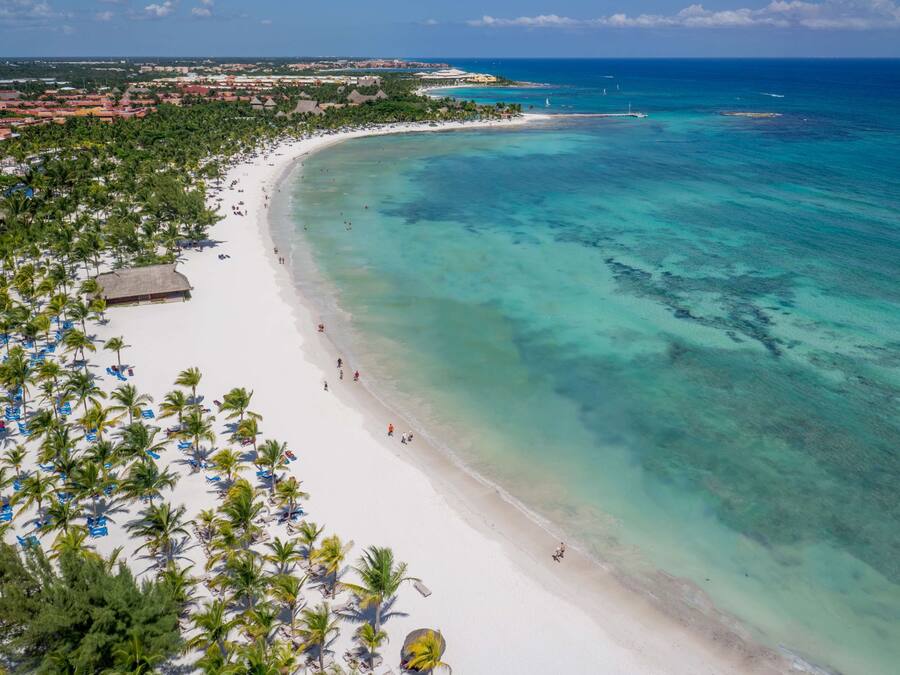 Una playa de arena blanca y agua turquesa cristalina. Palmeras y sombrillas se alinean a lo largo de la orilla.