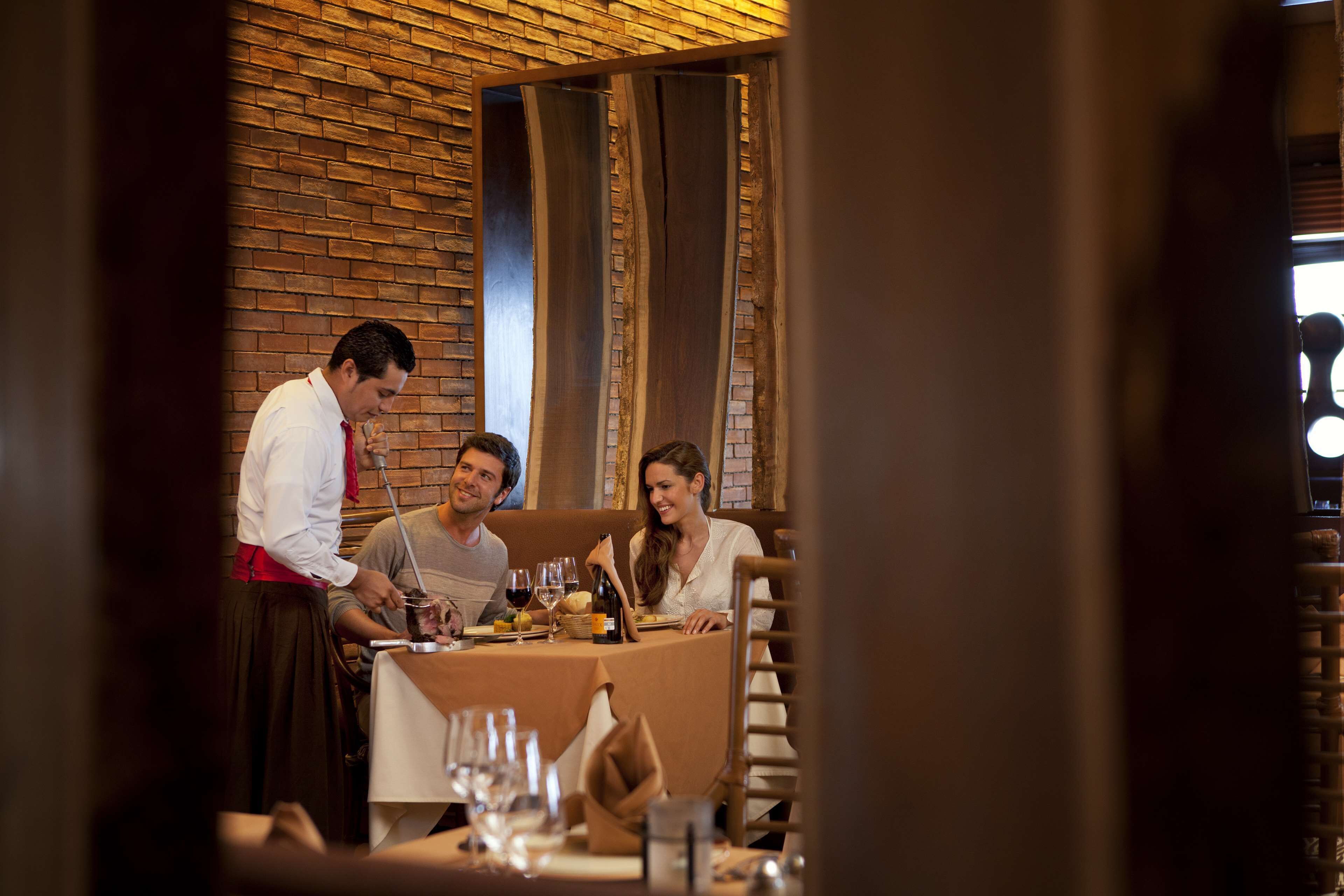 A restaurant with a brick wall and wooden beams, a table set with wine glasses and plates, and a waiter serving food.
