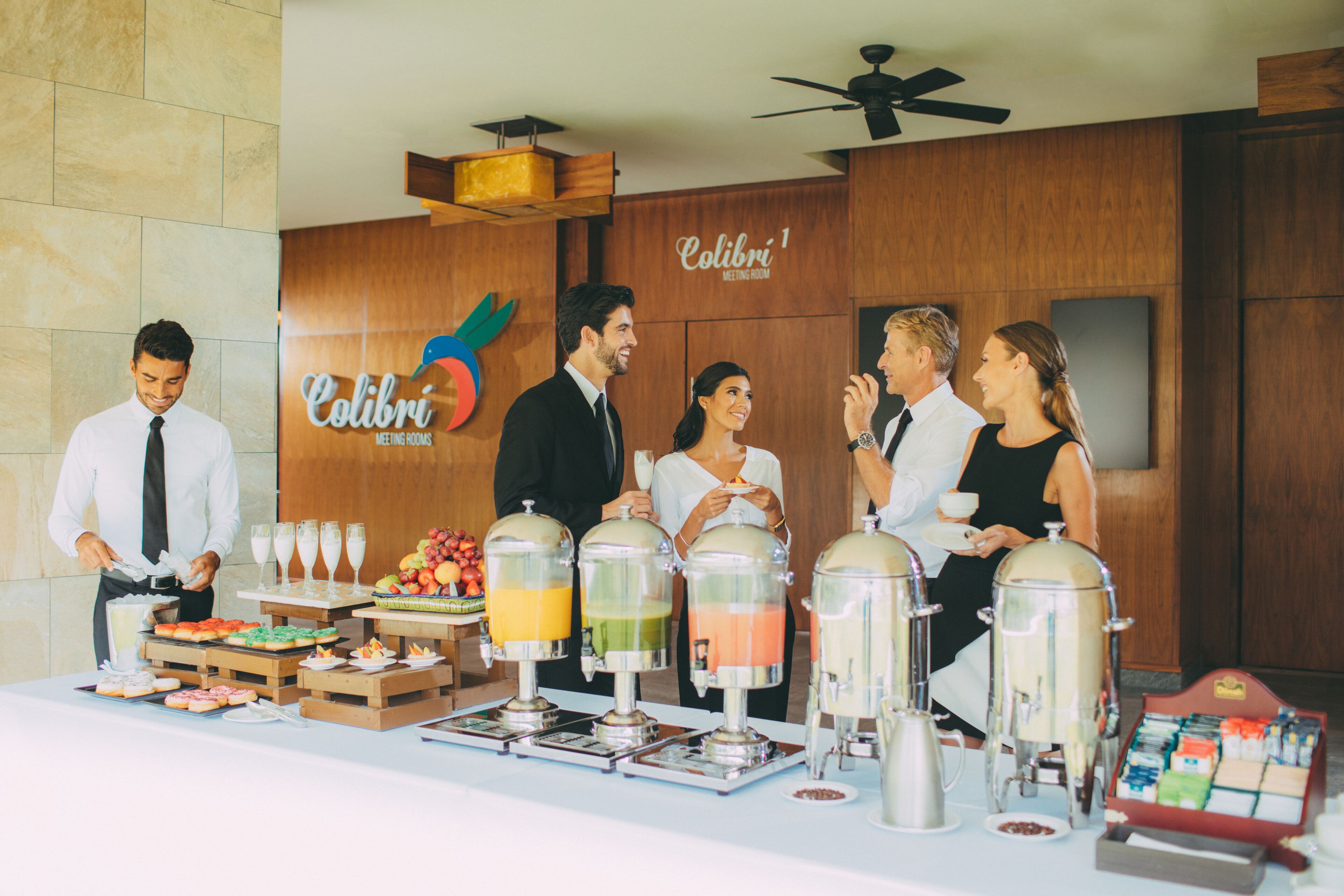 Ein Buffet mit verschiedenen Speisen und Getränkeautomaten, einem Schild mit der Aufschrift "Colibri Meeting Room" und einem Deckenventilator.