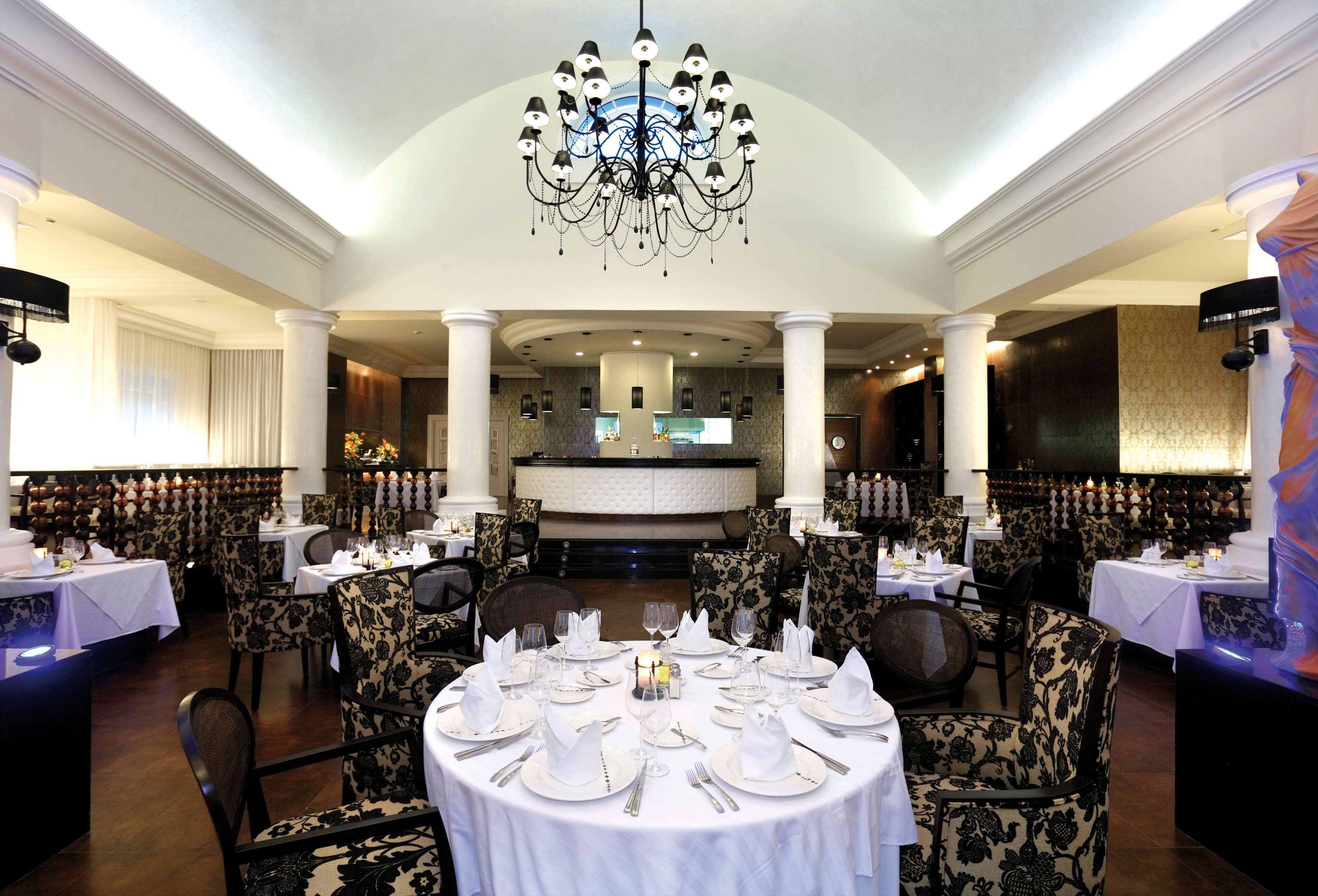 A formal dining area with round tables set for service, a central bar, and a chandelier.
