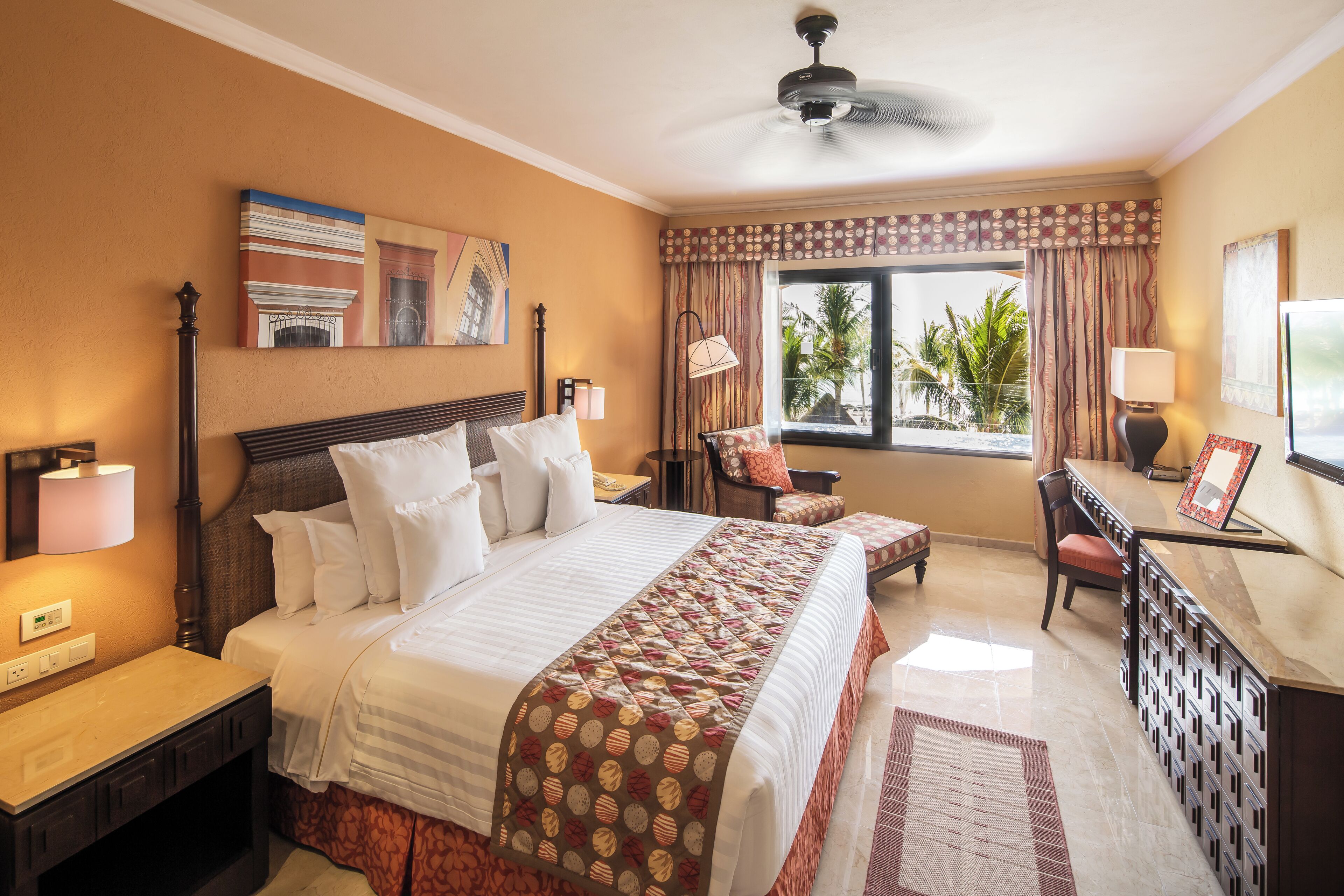 A hotel room with a large bed, a desk, a chair, a window with a view of palm trees, and a ceiling fan.