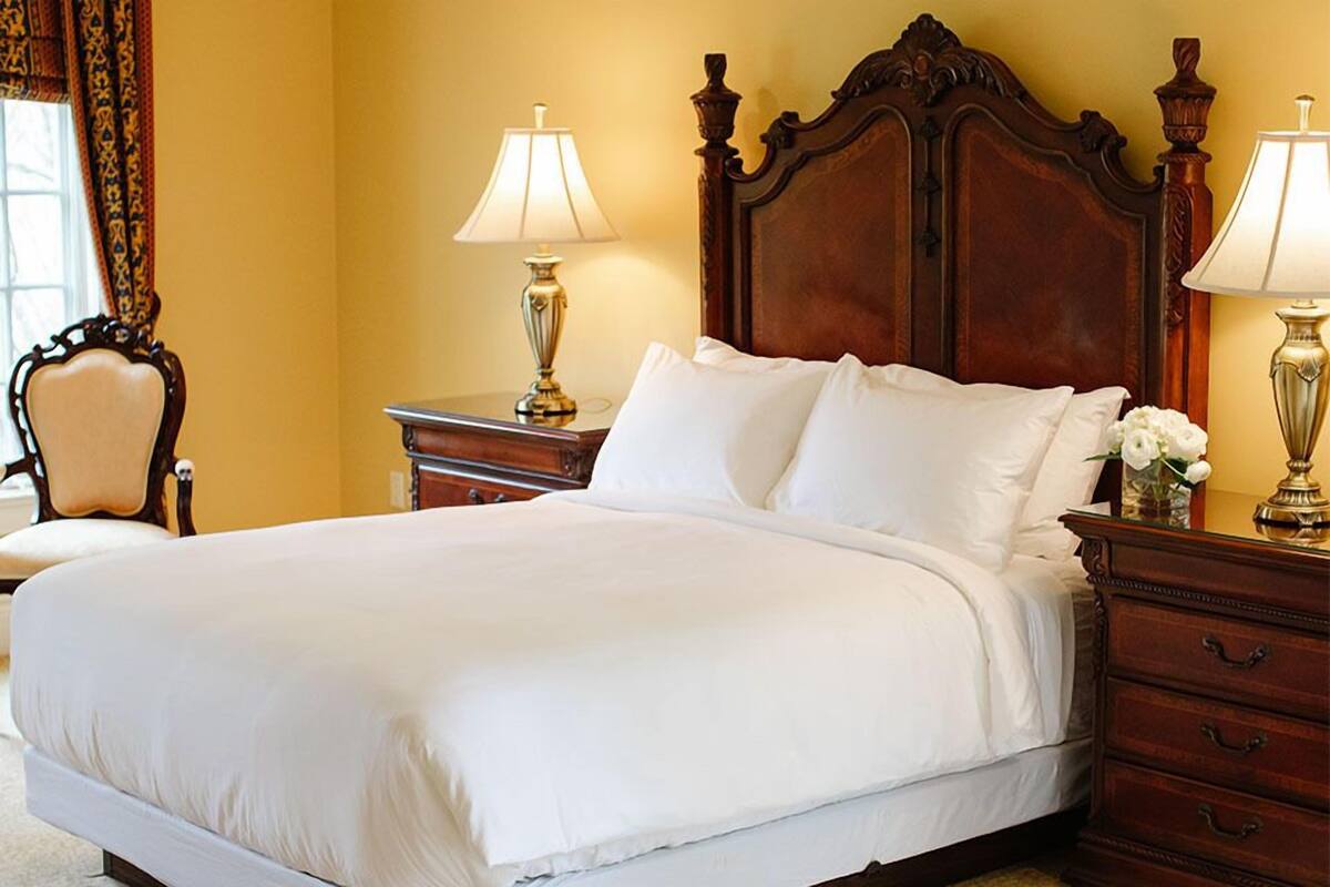 A large bed with white linens, a wooden headboard, two bedside tables with lamps, and a chair.