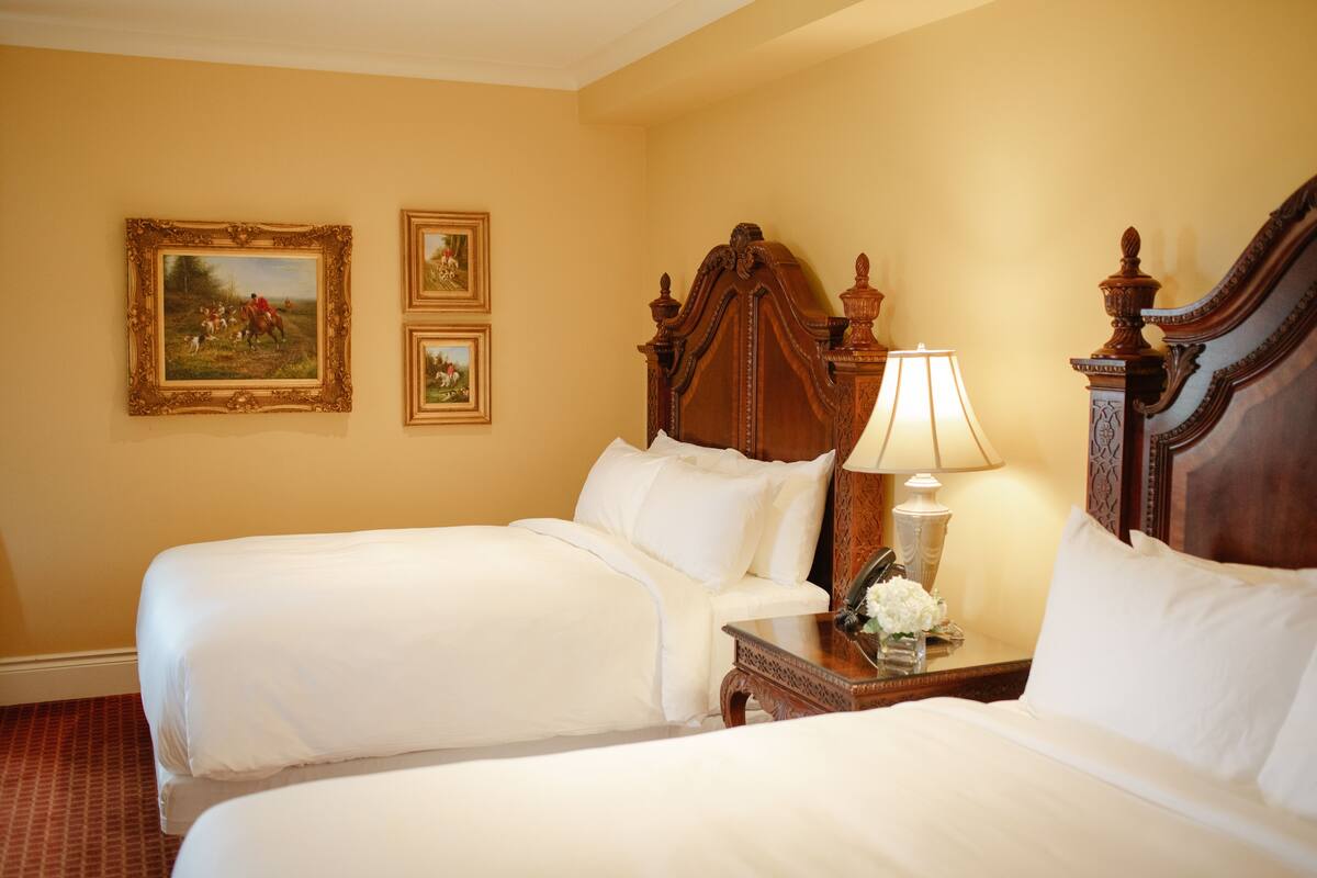 A hotel room with two beds, a wooden headboard, a bedside table with a lamp, and two framed pictures on the wall.