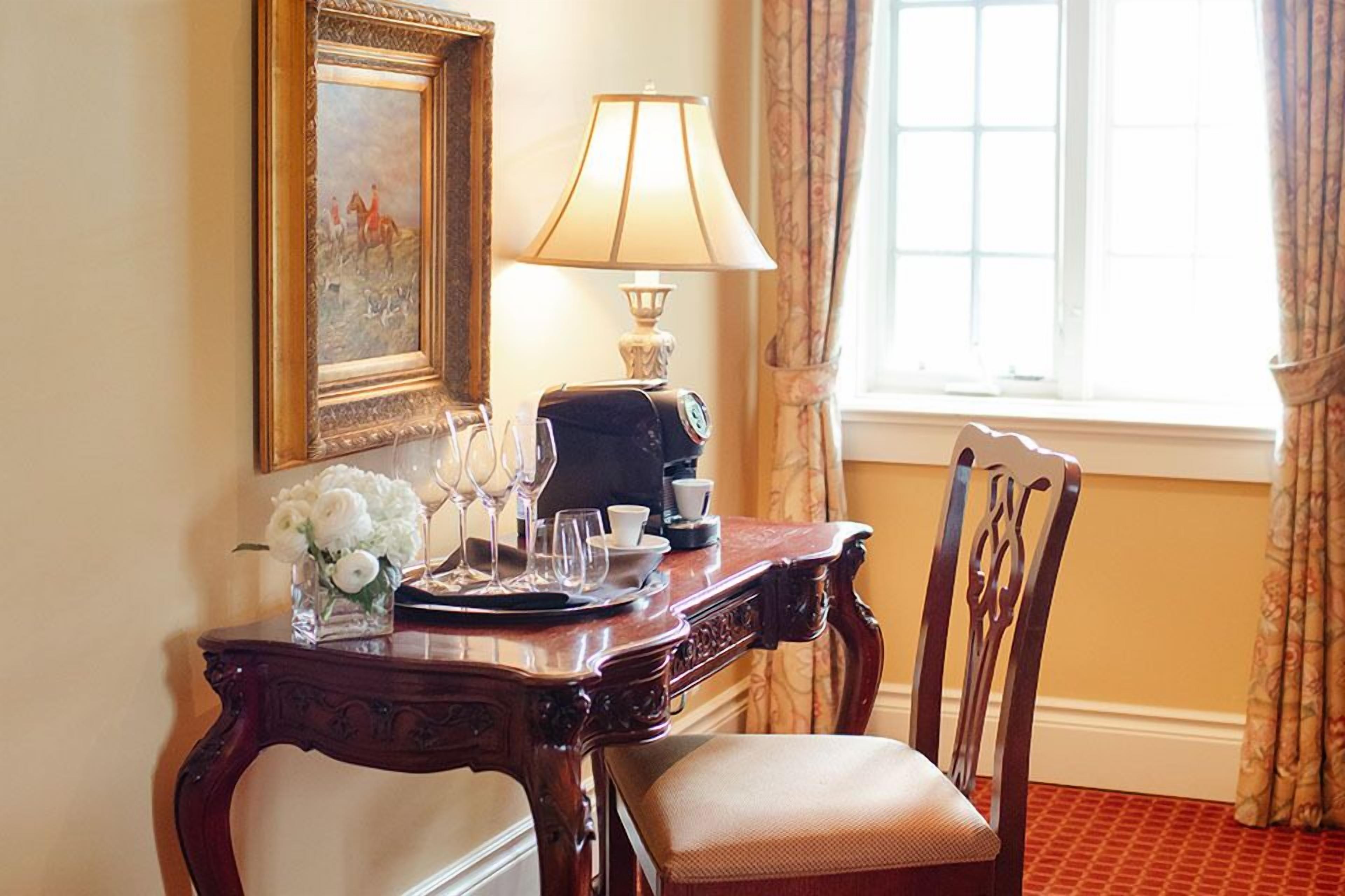 A hotel room with a wooden desk, a lamp, a coffee maker, a vase with flowers, and a chair.