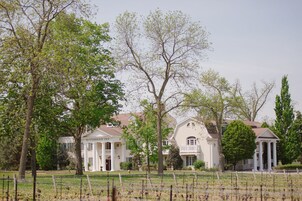 A large white building with a central pavilion, surrounded by trees and a fenced vineyard.