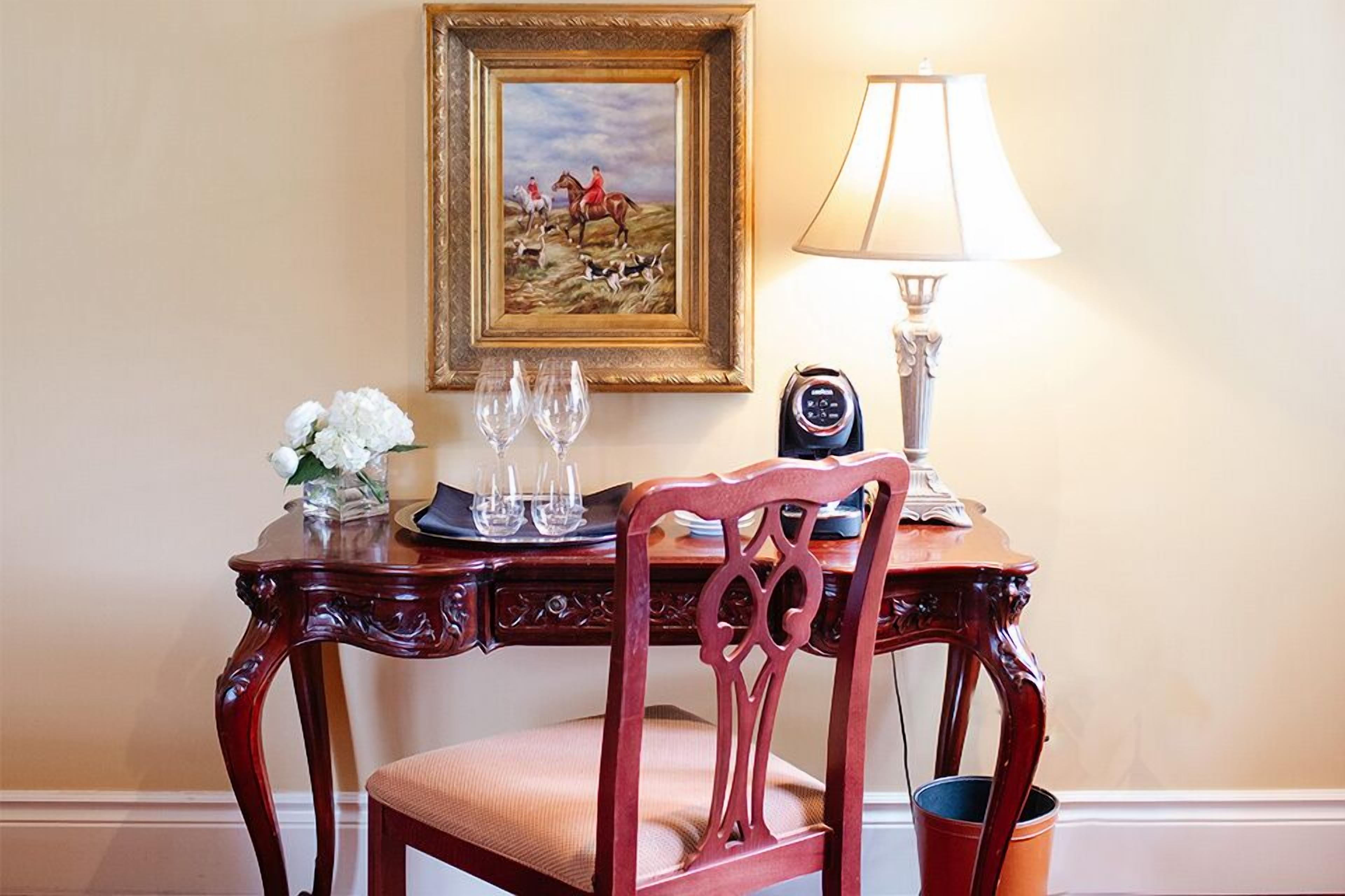 A wooden chair with a cushioned seat and backrest, positioned in front of a wooden table with a glass vase of white flowers, two wine glasses, and a tray. A framed painting of a horse race is on the wall above the table.