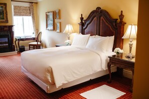 A hotel room with a large bed, two bedside tables with lamps, a chair, a fireplace, and framed pictures on the wall.