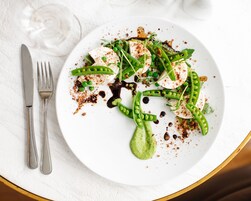 Un plato blanco con un guiso de verdura verde, un tenedor y un cuchillo sobre un mantel blanco.