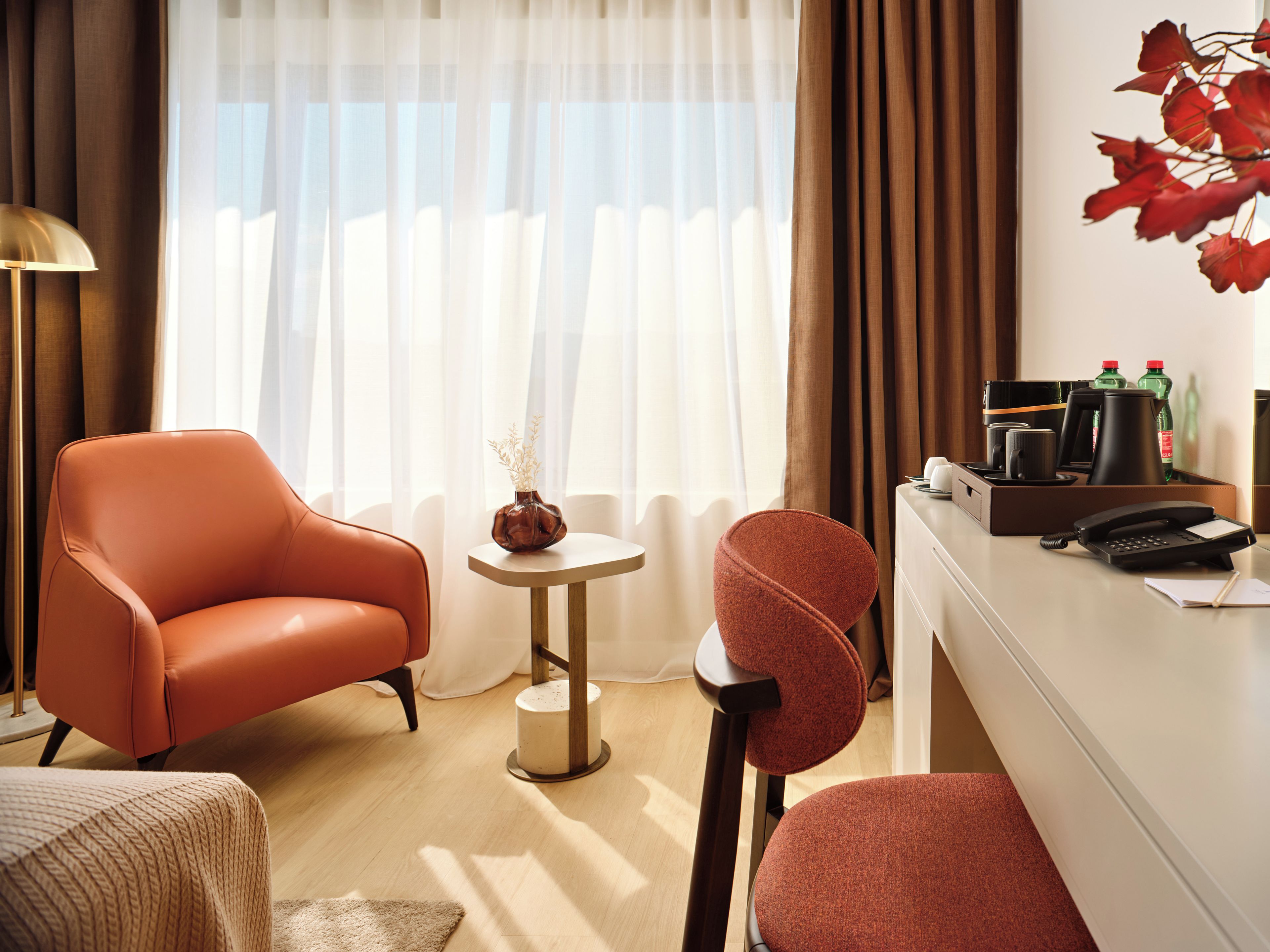 Habitación de hotel con un sillón de cuero, una mesita redonda con un florero, un escritorio con teléfono y cafetera, y vistas a través de cortinas traslúcidas.
