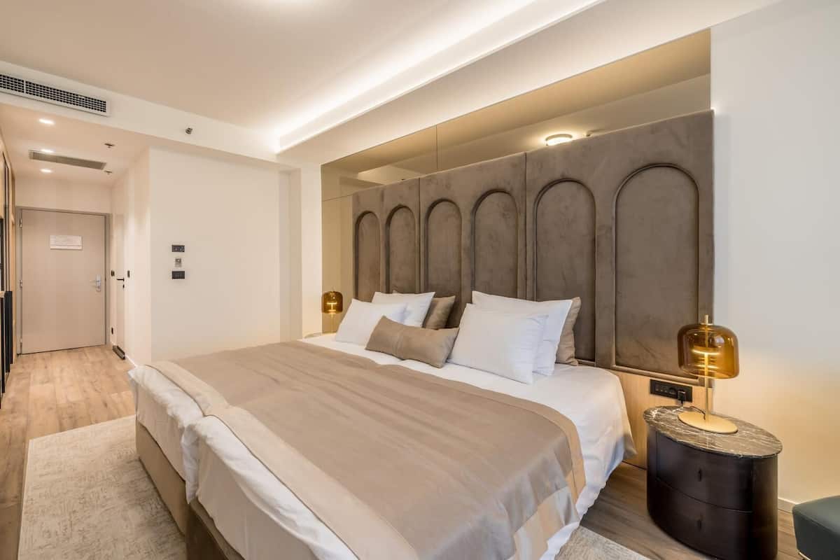 Una habitación de hotel moderna con una cama grande, cabecero con diseños circulares, mesita de noche con lámpara y puerta que da al pasillo.