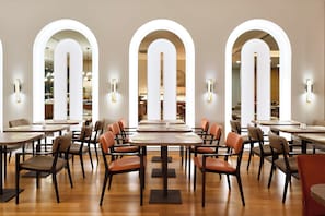 Un restaurante con varias mesas y sillas, ventanas arqueadas e iluminación instalada en la pared.