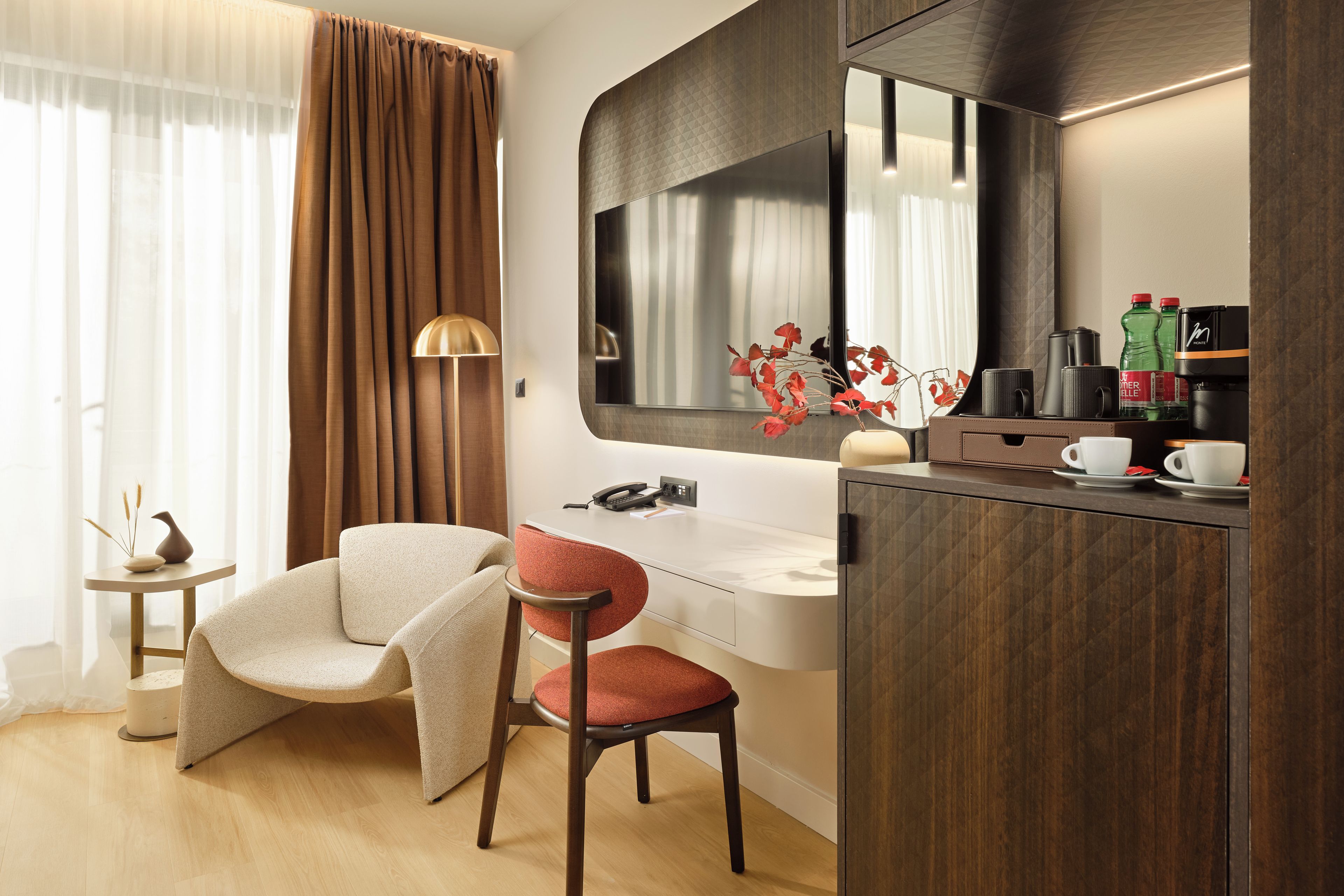 Ein modernes Hotelzimmer mit Holzboden, einem beigefarbenen Sessel, einem roten Stuhl, einem weißen Schreibtisch, einem Spiegel und einer Minibar.