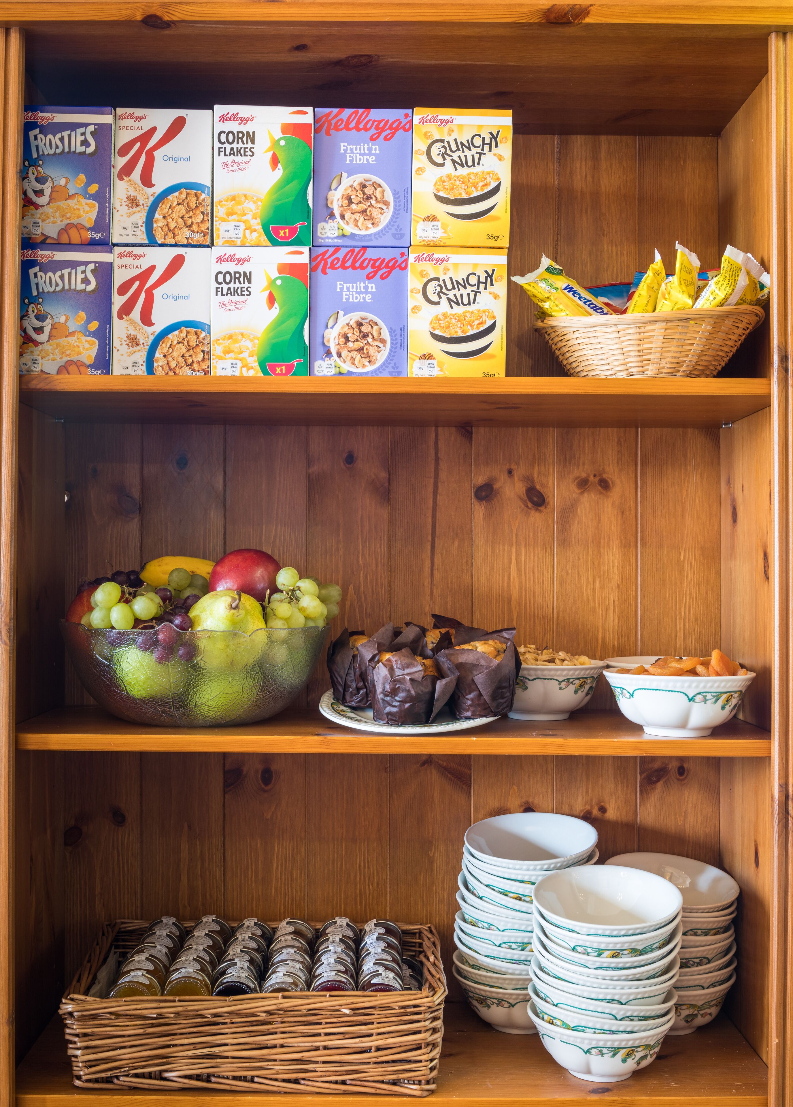 A wooden shelf with cereal boxes, a fruit bowl, wrapped pastries, and stacked bowls.