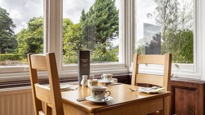 Una mesa para el desayuno dispuesta con tazas y platillos, un atril para el menú, y una vista de árboles y un edificio en el exterior.