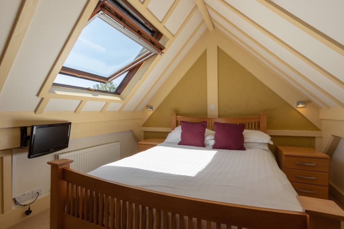 A bedroom with a sloped ceiling, a bed with white bedding and purple pillows, a bedside table, a TV mounted on the wall, and a window with a view of the sky.