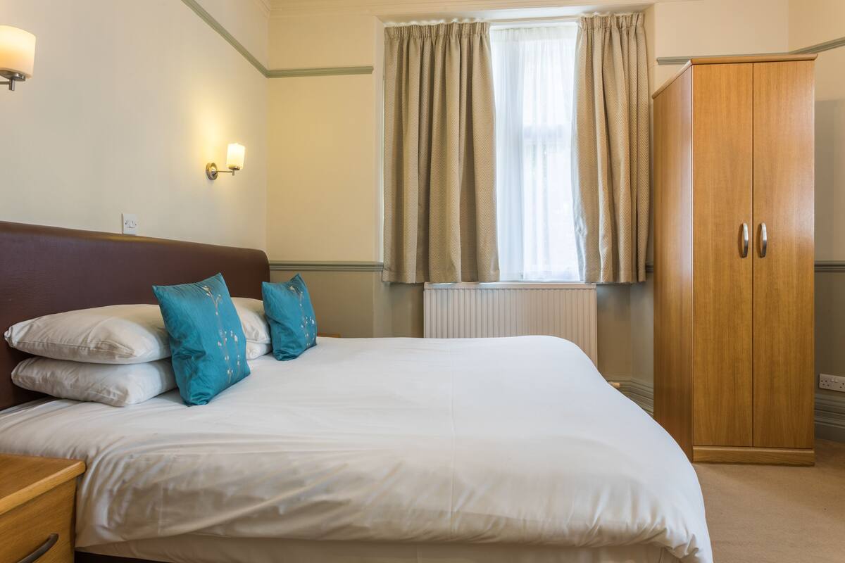 A neatly made bed with white and blue pillows, a wooden wardrobe, and a window with curtains.