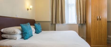 Una cama bien hecha con almohadas blancas y azules, un armario de madera y una ventana con cortinas.