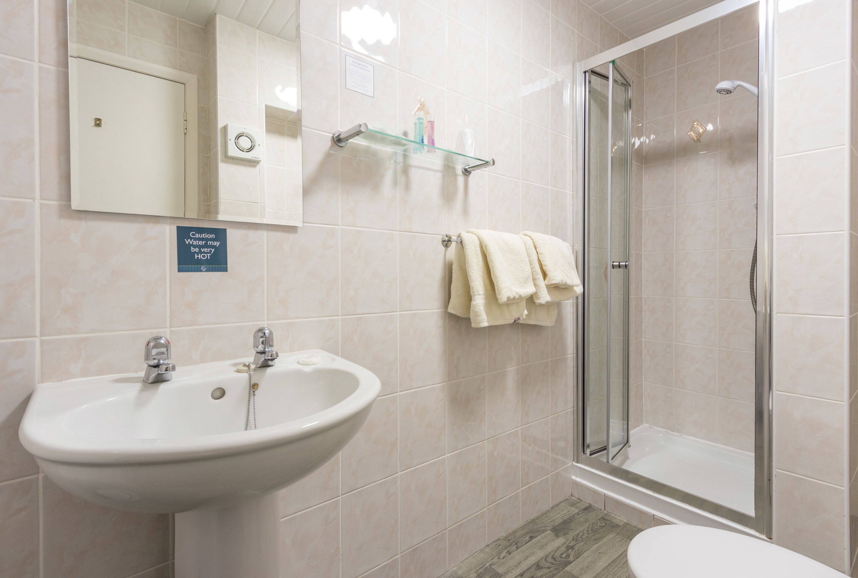 A bathroom with a sink, mirror, glass shower enclosure, and a caution sign about hot water.