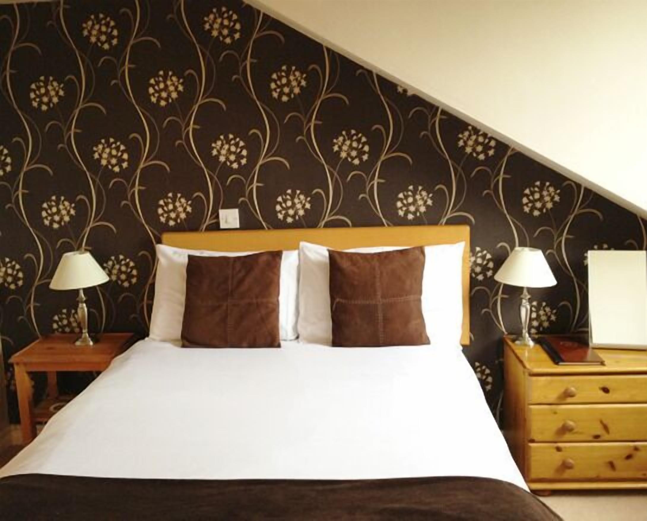 A hotel room with a large bed, two bedside tables with lamps, a wooden nightstand, and a patterned wallpaper.