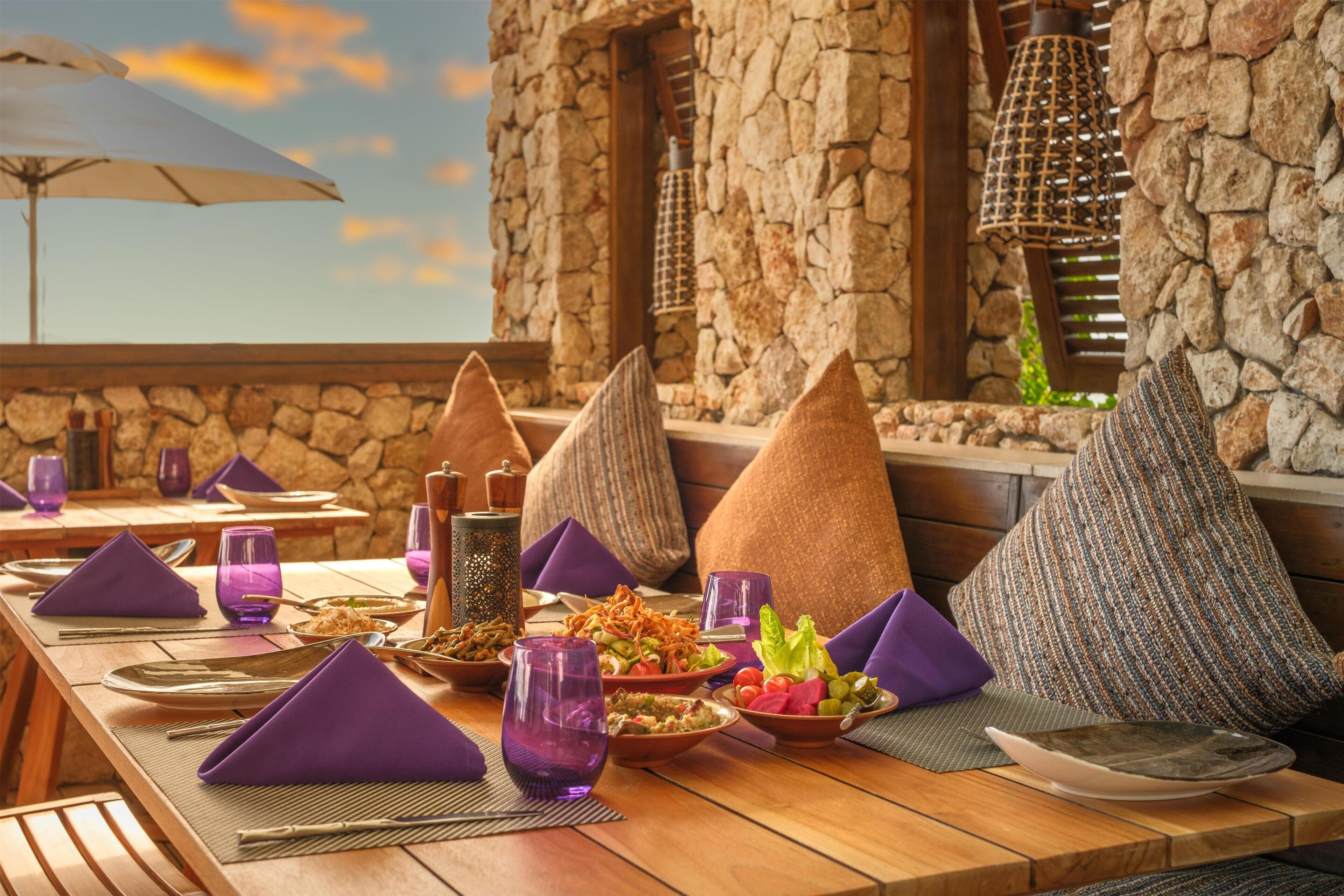 A wooden table set with purple napkins, plates, and glasses, with stone walls and a view of the sky.