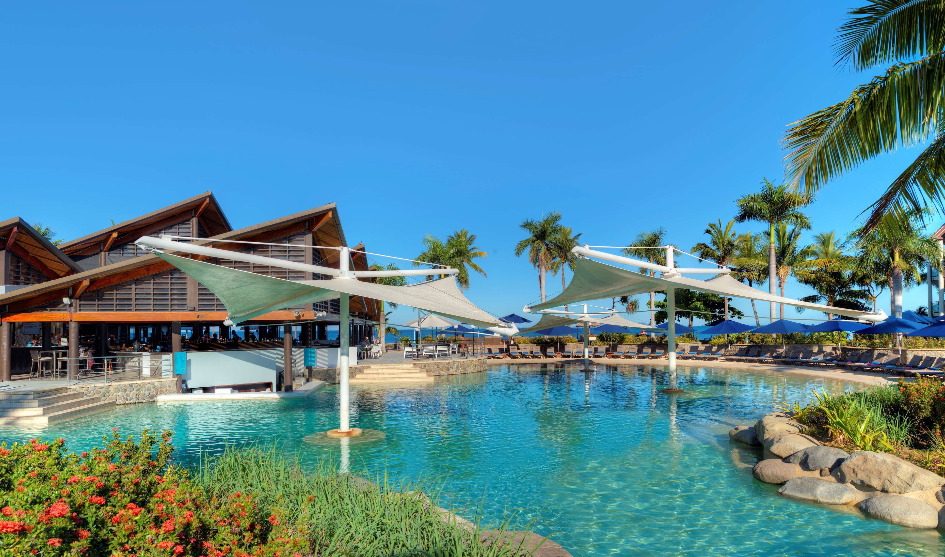 Un espace piscine aménagé dans un complexe hôtelier, avec des chaises longues, des parasols et un bâtiment à la façade en bois.