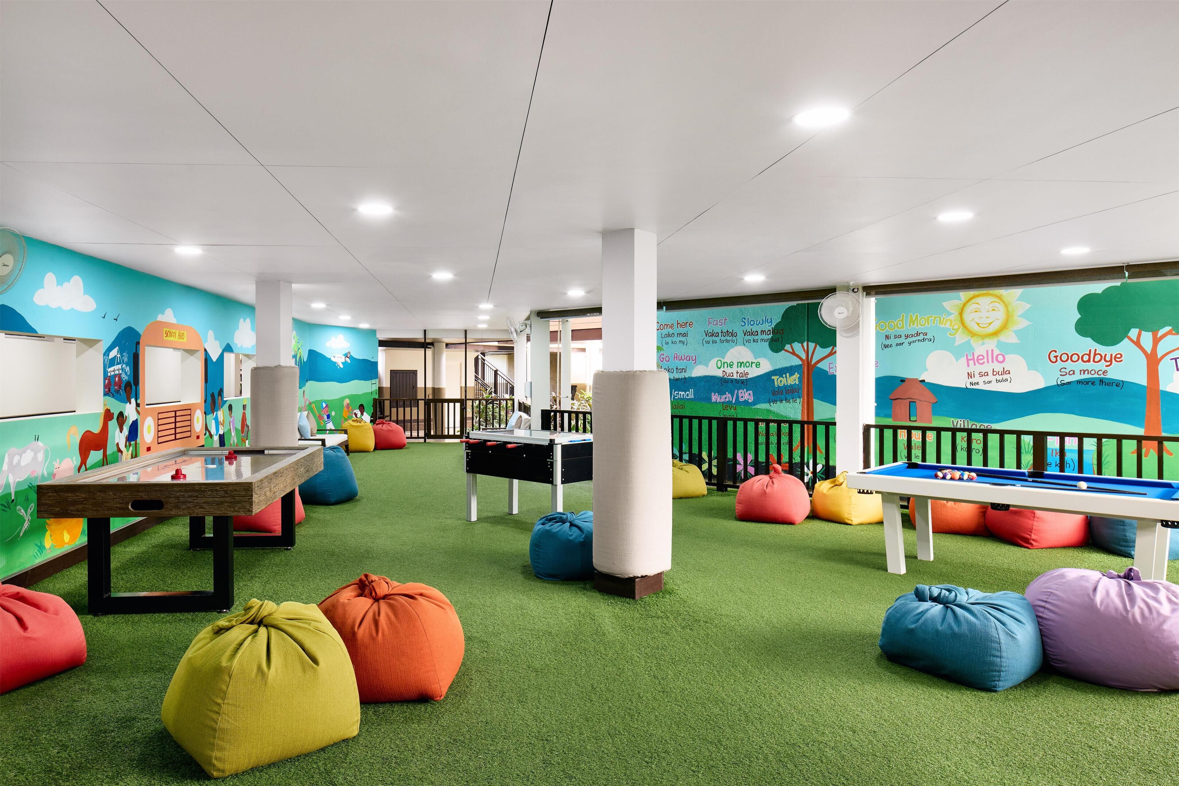 Zona de juegos para niños con césped artificial verde, pufs de colores, mesa de billar y dardos.