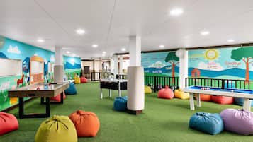 Zona de juegos para niños con césped artificial verde, pufs de colores, mesa de billar y dardos.