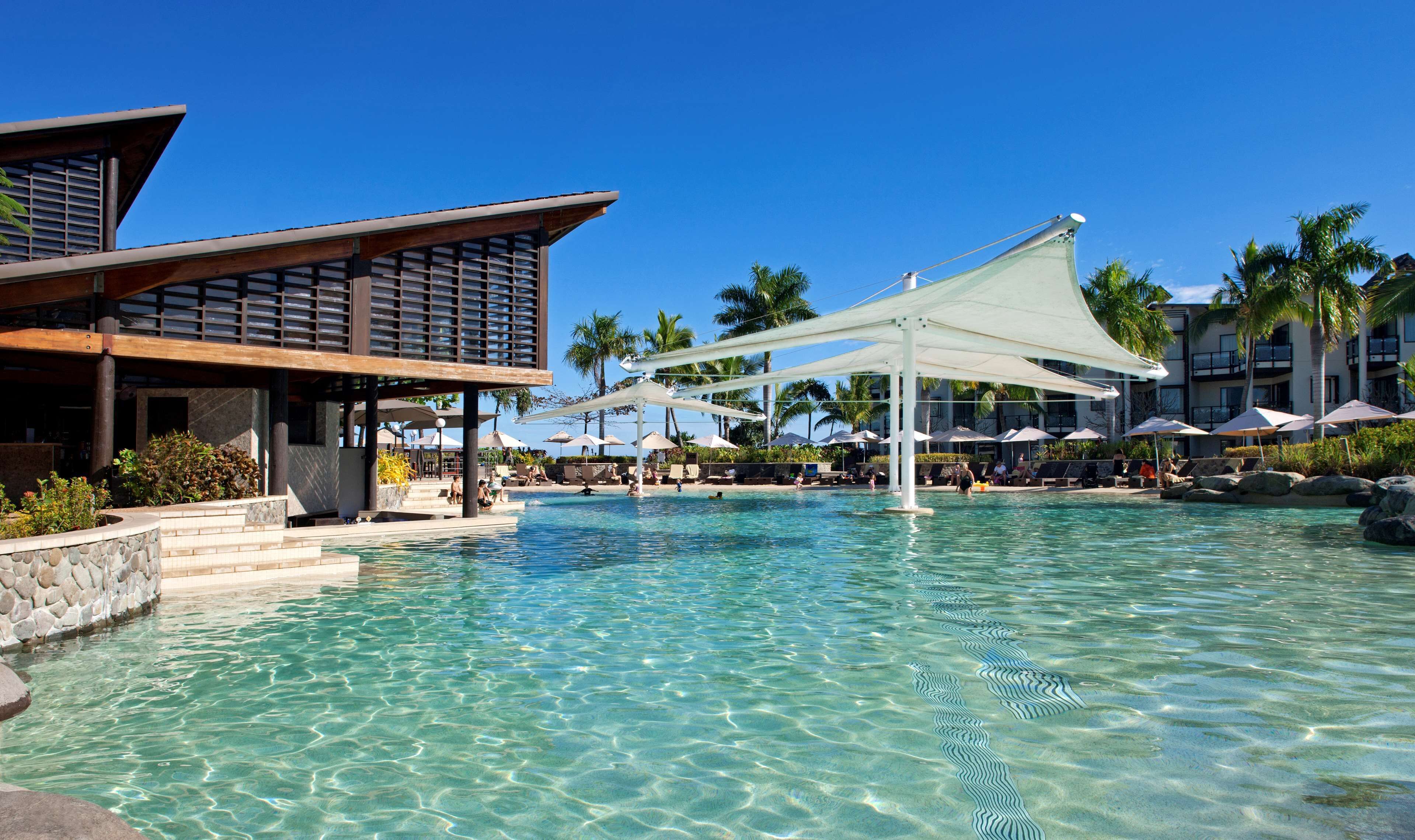 Una piscina nítida amb una estructura de protecció solar, seients de relax i un edifici amb teulada inclinada.