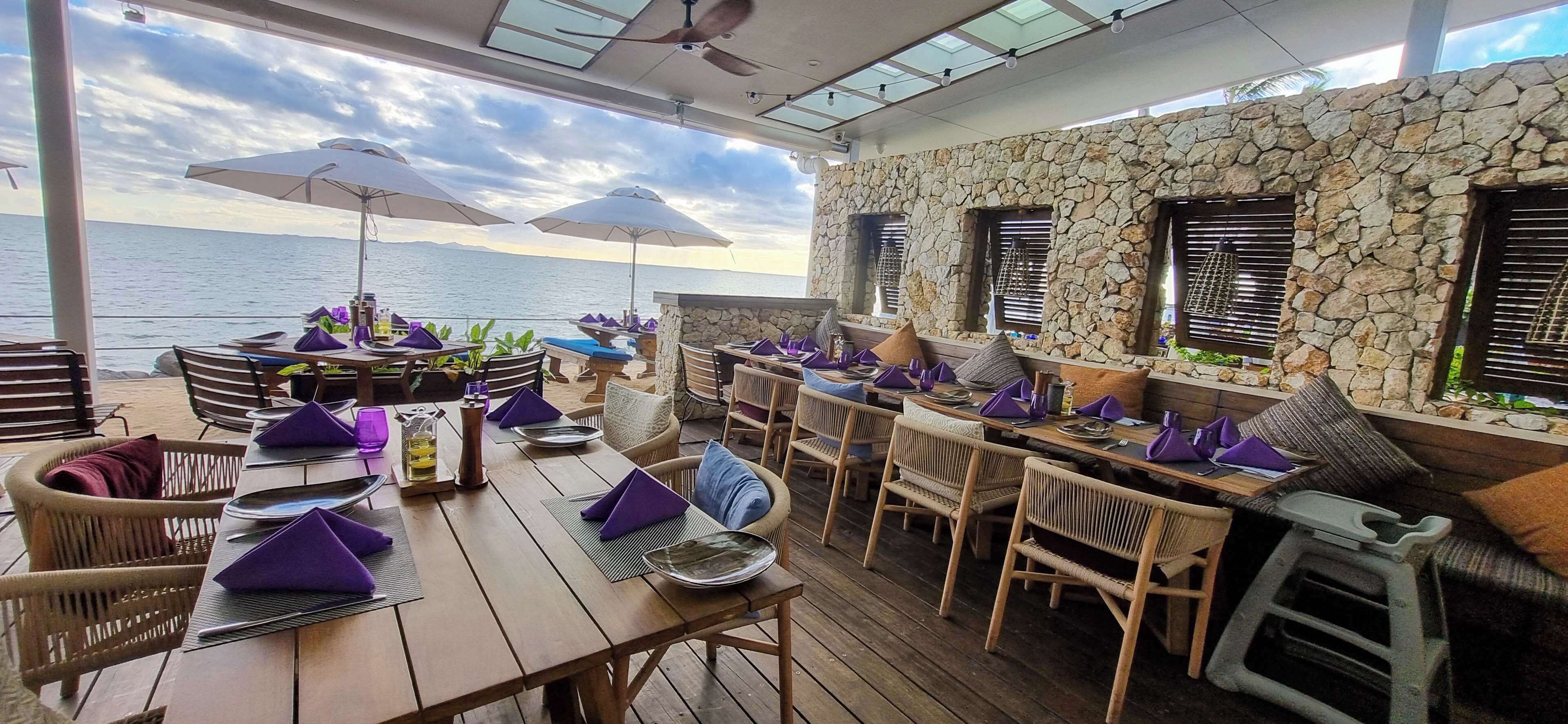 Un área de comedor al aire libre con mesas y sillas de madera, paredes de piedra y sombrillas.