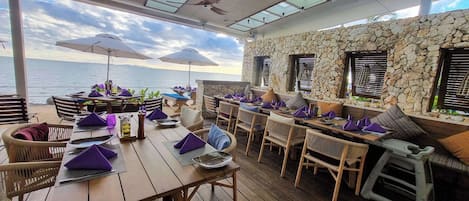 Un área de comedor al aire libre con mesas y sillas de madera, paredes de piedra y sombrillas.