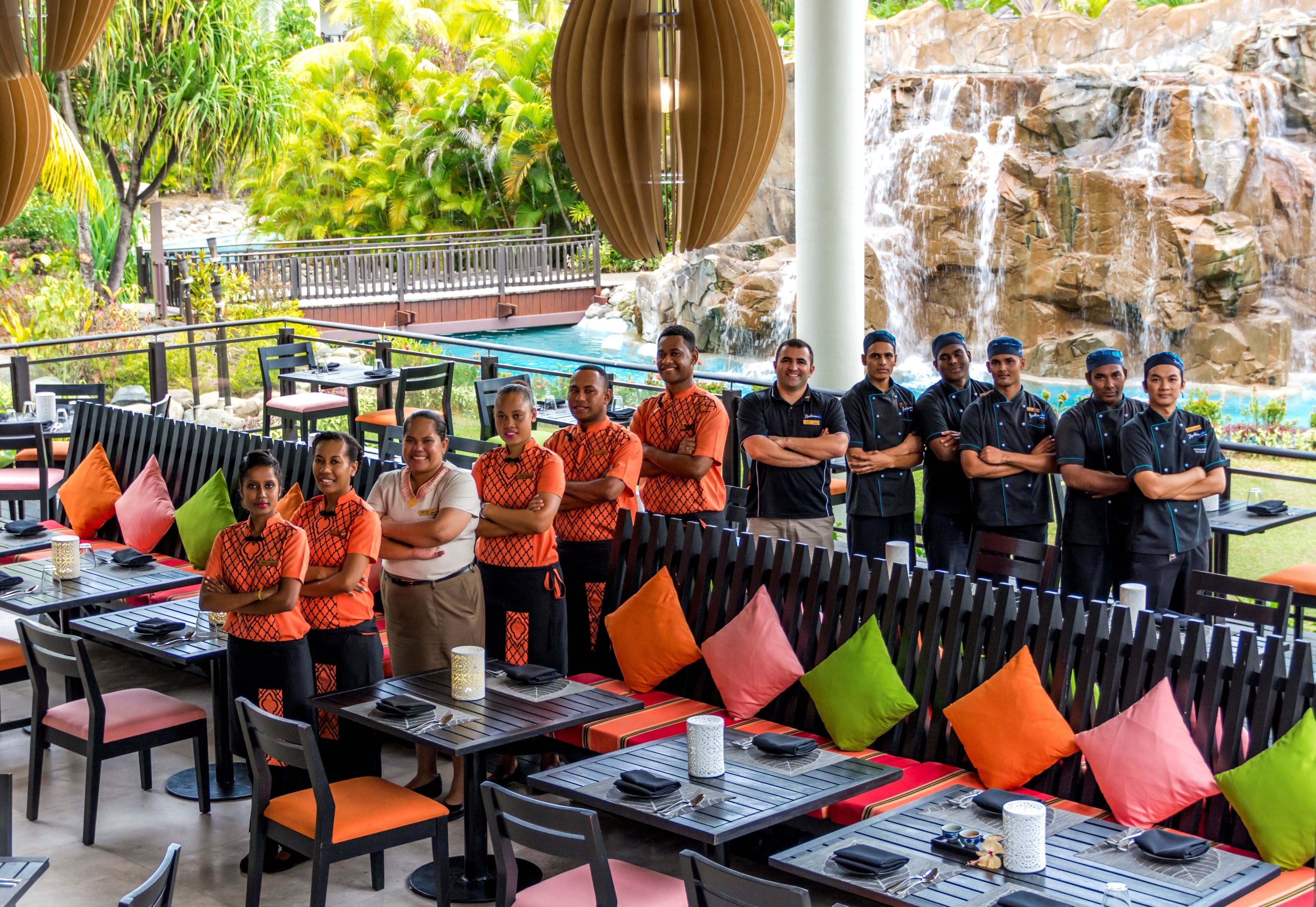A group of people standing in an outdoor dining area with tables and chairs, a pool, and a waterfall in the background.