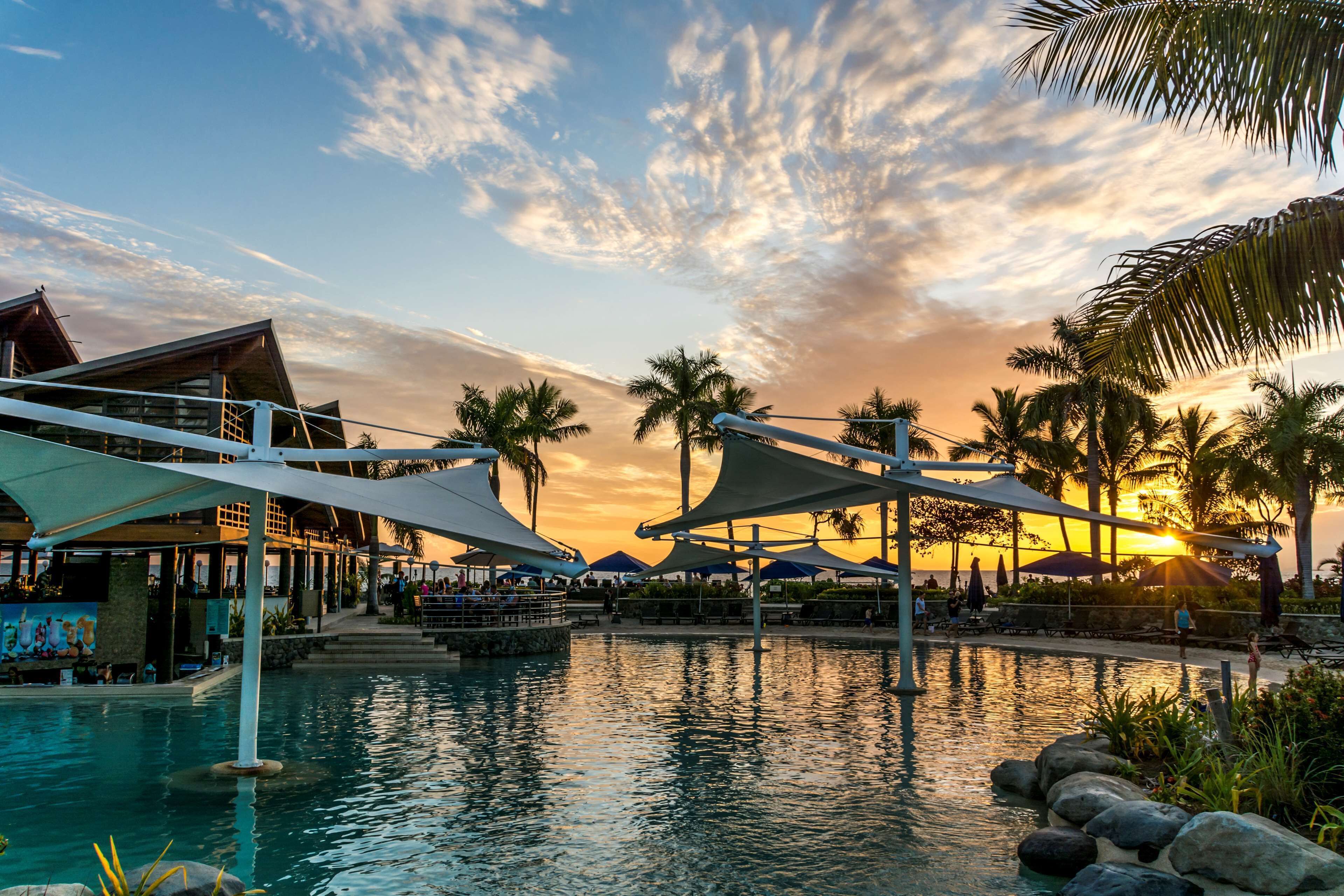 Ein Poolbereich eines Resorts mit Sonnenuntergangsblick, Palmen und schattigen Sitzbereichen.