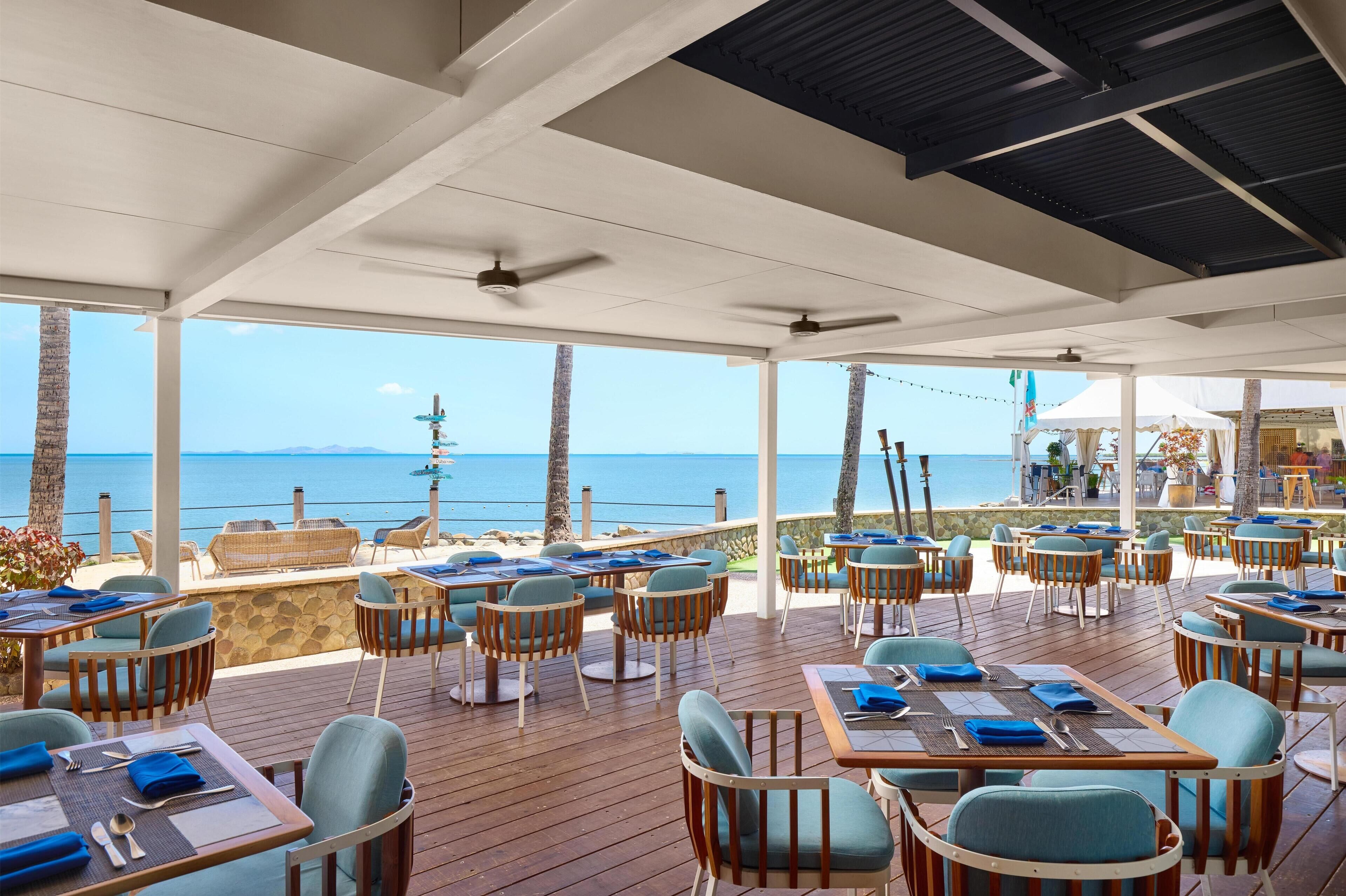 An outdoor dining area with tables and chairs, overlooking a beach and the sea.