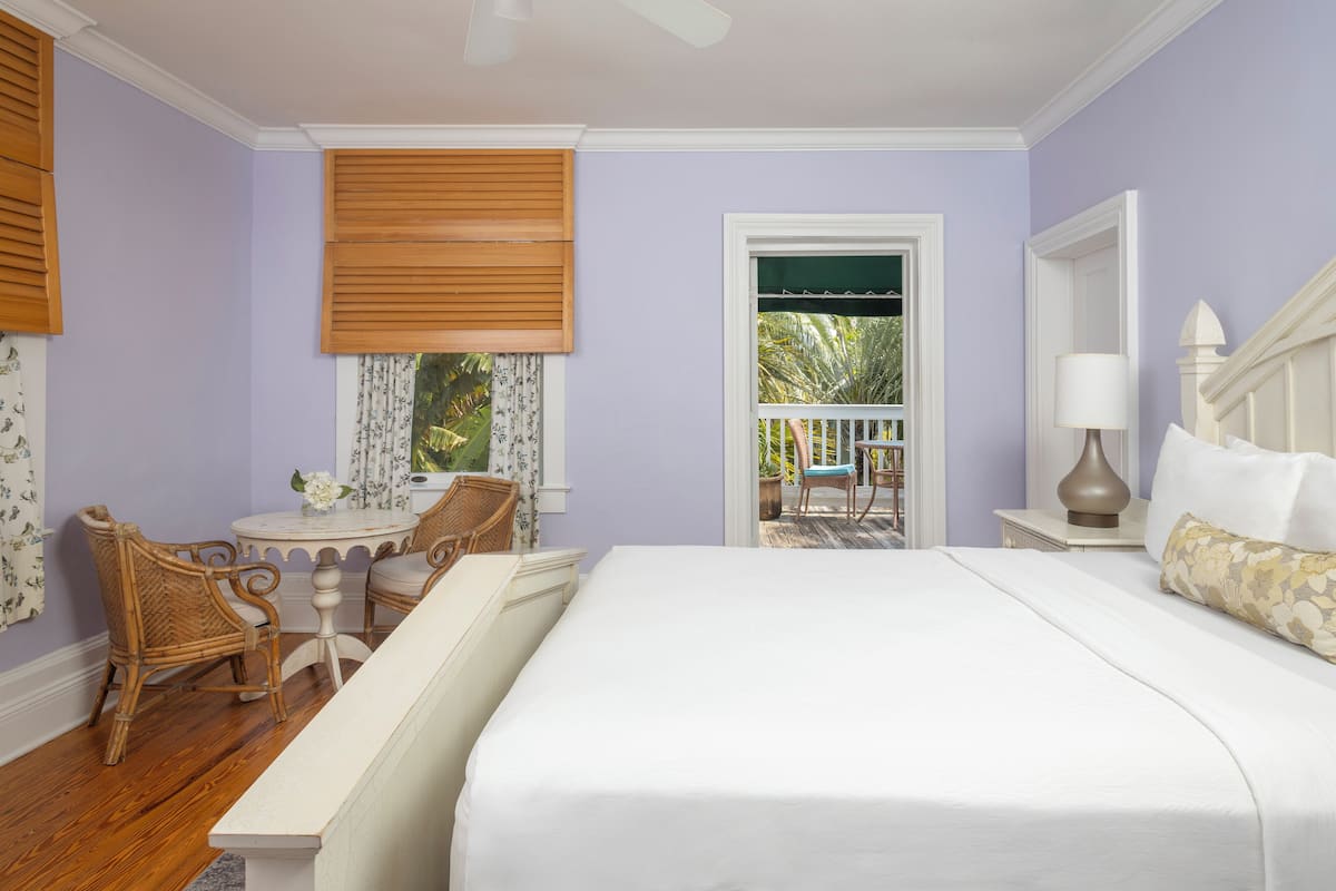 A bedroom with a large bed, a small table, two chairs, a window with blinds, and a view of the outdoors.